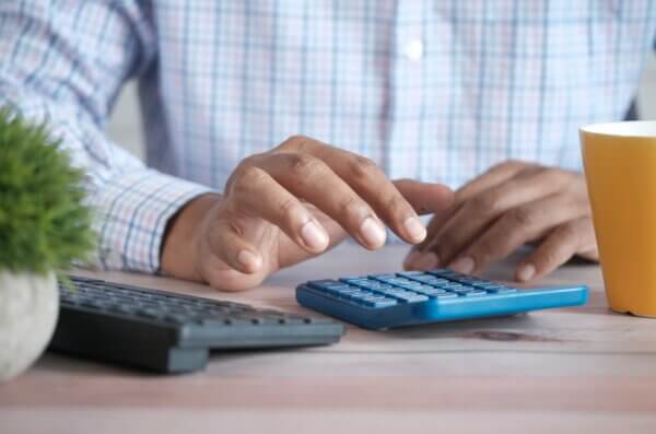 mężczyzna dokonuje obliczeń na kalkulatorze, obok stoją żółty kubek i klawiatura
